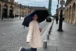 Woman in a cream coat with black umbrella on a street.