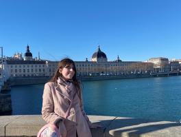 Woman in a light brown coat in front of a castle over water.