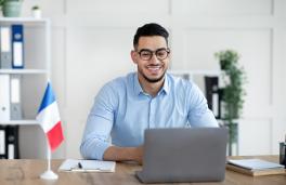 Man with glasses looking at a laptop with a French flag in the background.