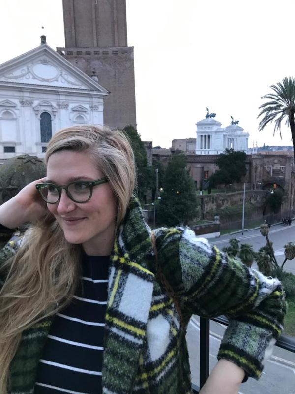 Photograph of a white woman with long blonde hair, wearing green glasses, a blue-and-white striped sweater, and a green-and-white tartan coat, standing with a view of Rome in the background (visible is the Church of Santa Caterina a Magnanapoli and the Monument to Vittorio Emanuele II)